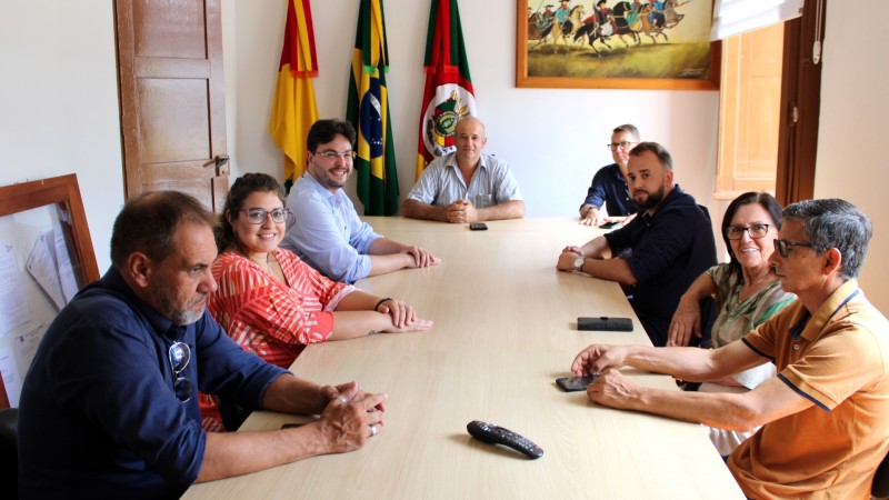 Em traje formal, os representantes do Procon RS e do Procon Municipal de Piratini estão sentados ao redor de uma mesa comprida e retangular. Ao fundo, estão hasteadas as bandeiras do Brasil, Rio Grande do Sul e do município de Piratini.
