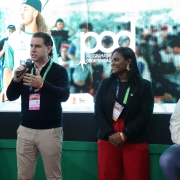 Há três pessoas em um palco. Ao fundo um painel de apresentação. Um homem está de pé e segurando um microfone. Uma mulher, ao lado dele, olha para ele. E outro homem está sentado.