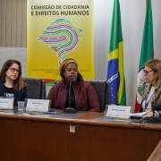 Mulher negra, haitiana, com casaco vermelho, blusa preta, gorro amarelo. Sentada, falando ao microfone.