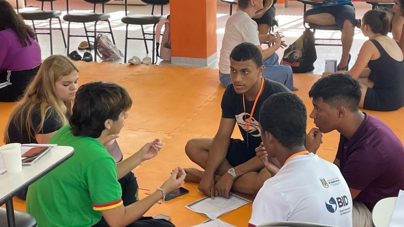 A imagem mostra um grupo de cinco jovens sentados no ch&atilde;o, em c&iacute;rculo, aparentemente participando de uma atividade em grupo ou discuss&atilde;o. Eles est&atilde;o em um espa&ccedil;o interno amplo com piso laranja, que lembra um ambiente de evento, workshop ou din&acirc;mica de grupo. No centro do grupo h&aacute; pap&eacute;is e um celular, sugerindo que est&atilde;o trabalhando em alguma tarefa ou projeto juntos. Um dos jovens, vestindo uma camiseta verde, parece estar explicando algo enquanto gesticula com as m&atilde;os. Os outros quatro est&atilde;o atentos, olhando para ele ou para o centro da roda, indicando que est&atilde;o ouvindo ou debatendo ideias. Alguns deles usam crach&aacute;s pendurados no pesco&ccedil;o, o que refor&ccedil;a a ideia de que participam de um evento organizado, como um curso, treinamento ou encontro estudantil. Ao fundo, h&aacute; outras pessoas tamb&eacute;m sentadas e conversando em pequenos grupos, al&eacute;m de cadeiras e mesas, mostrando que v&aacute;rias atividades semelhantes acontecem no mesmo espa&ccedil;o.