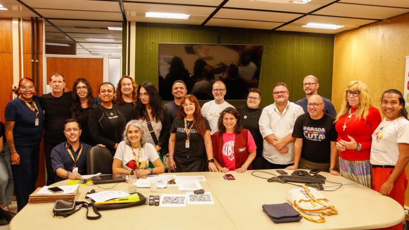 Imagem em sala de reuni&otilde;es ampla e bem iluminada, com paredes de madeira clara e painel verde ao fundo. H&aacute; um grupo numeroso de adultos posando para a foto, 11 mulheres e 7 homens, s&atilde;o pessoas da comiss&atilde;o eleitoral e novos conselheiros e conselheiras eleitas, ao redor de uma mesa clara grande. Algumas pessoas est&atilde;o em p&eacute; e outras sentadas &agrave; frente. Sobre a mesa h&aacute; pap&eacute;is, cadernos, bolsas, celulares e um leitor de QR Code para a vota&ccedil;&atilde;o que havia acontecido. Todas usam roupas casuais e crach&aacute;s. Ao fundo, h&aacute; uma tela preta desligada. Momento final ap&oacute;s o an&uacute;ncio dos eleitos e eleitas.