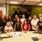 Imagem em sala de reuni&otilde;es ampla e bem iluminada, com paredes de madeira clara e painel verde ao fundo. H&aacute; um grupo numeroso de adultos posando para a foto, 11 mulheres e 7 homens, s&atilde;o pessoas da comiss&atilde;o eleitoral e novos conselheiros e conselheiras eleitas, ao redor de uma mesa clara grande. Algumas pessoas est&atilde;o em p&eacute; e outras sentadas &agrave; frente. Sobre a mesa h&aacute; pap&eacute;is, cadernos, bolsas, celulares e um leitor de QR Code para a vota&ccedil;&atilde;o que havia acontecido. Todas usam roupas casuais e crach&aacute;s. Ao fundo, h&aacute; uma tela preta desligada. Momento final ap&oacute;s o an&uacute;ncio dos eleitos e eleitas.