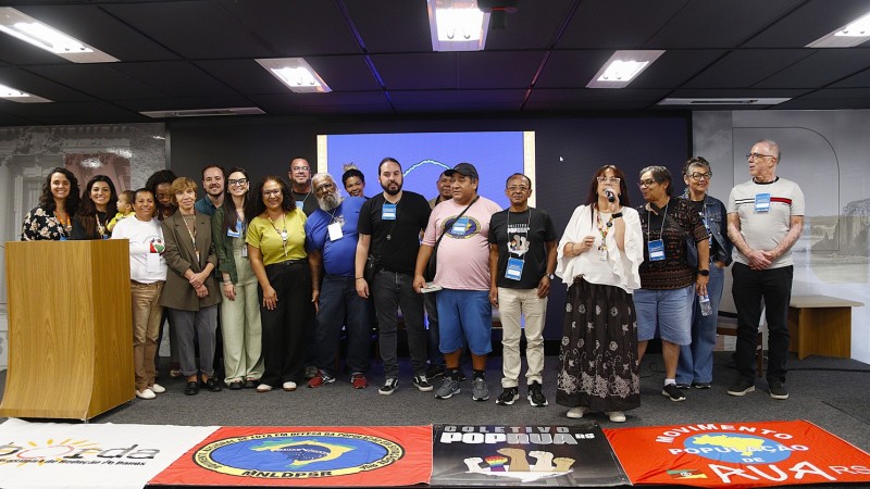 Grupo de pessoas em p&eacute;, em audit&oacute;rio, uma falando ao microfone. No ch&atilde;o, bandeiras de movimentos sociais ligadas a entidades voltadas &agrave;s pessoas em situa&ccedil;&atilde;o de rua.