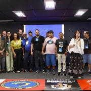 Grupo de pessoas em p&eacute;, em audit&oacute;rio, uma falando ao microfone. No ch&atilde;o, bandeiras de movimentos sociais ligadas a entidades voltadas &agrave;s pessoas em situa&ccedil;&atilde;o de rua.