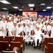 Foto de um grande grupo de pessoas reunidas em um audit&oacute;rio, organizadas em v&aacute;rias fileiras, de p&eacute;, voltadas para a c&acirc;mera. Muitas seguram certificados retangulares com moldura clara. As roupas s&atilde;o variadas, com predomin&acirc;ncia de branco e estampas tradicionais. &Agrave; frente, h&aacute; cadeiras de madeira vazias. Ao fundo, um painel com fundo colorido e textos indica um evento institucional, sob ilumina&ccedil;&atilde;o de teto uniforme.