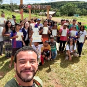 Foto ao ar livre, em &aacute;rea rural. Em primeiro plano, um homem negro, servidor da SJCDH, segura o celular e tira uma selfie. Ao fundo, um grande grupo de adultos e crian&ccedil;as ind&iacute;genas est&aacute; reunido sobre a grama, voltados para a c&acirc;mera, alguns em p&eacute; e outros agachados, muitos segurando documentos claros, suas certid&otilde;es de nascimento. Atr&aacute;s do grupo h&aacute; uma estrutura de madeira com a frase &ldquo;XEE &hearts; TEKO&Aacute; POR&Atilde;&rdquo;. O cen&aacute;rio inclui cercas, &aacute;rvores, morros verdes e algumas constru&ccedil;&otilde;es simples.