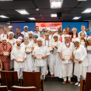 Fotografia mostra um grande grupo de cerca de trinta pessoas adultas reunidas em um auditório, algumas em pé sobre o palco e outras nas primeiras fileiras, todas voltadas para a câmera. Muitas seguram certificados retangulares com moldura clara à frente do corpo. As vestimentas são variadas, misturando roupas claras, peças estampadas, trajes sociais e vestimentas tradicionais com colares, lenços e turbantes. Ao fundo há um painel digital vermelho com textos em branco. À frente aparecem cadeiras de madeira do auditório. O piso é cinza e a iluminação é uniforme.