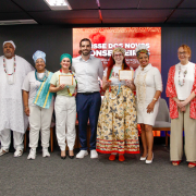 Fotografia mostra um grupo de nove pessoas adultas em pé, alinhadas em um palco de ambiente interno. Todas estão voltadas para a câmera; quatro seguram certificados emoldurados com as duas mãos. As roupas são variadas: há trajes sociais, roupas claras, peças estampadas e vestimentas tradicionais com tecidos longos, colares e turbantes. Ao fundo, um grande painel digital vermelho exibe textos em branco e grafismos. O piso é cinza, há cadeiras vazias atrás do grupo e a iluminação é uniforme.