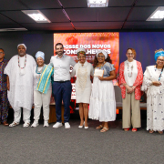 Fotografia mostra um grupo de doze pessoas adultas em pé, alinhadas sobre um palco em ambiente interno. Todas estão voltadas para a câmera; quatro seguram certificados retangulares com as duas mãos, à frente do corpo. As vestimentas são diversas, incluindo roupas sociais, peças claras, tecidos estampados e trajes tradicionais com colares, turbantes e panos longos. Ao fundo há um grande painel digital vermelho com textos e grafismos em branco.