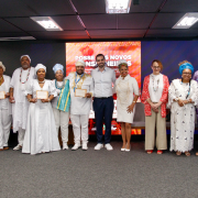 Fotografia mostra um grupo de cerca de doze pessoas adultas em pé, alinhadas lado a lado sobre um palco em um auditório. Todas estão voltadas para a câmera. A maioria segura certificados retangulares com moldura clara, posicionados à frente do corpo. As vestimentas são variadas, com roupas sociais, peças claras, tecidos coloridos e trajes tradicionais com colares, turbantes e panos longos. Ao fundo há um grande painel digital vermelho com textos e grafismos em branco. O piso é cinza, o teto possui luminárias retangulares e há cadeiras dispostas atrás do grupo.