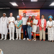 Fotografia mostra um grupo de cerca de doze pessoas adultas em pé, lado a lado, sobre um palco em um auditório. Todas estão voltadas para a câmera; várias seguram certificados emoldurados à frente do corpo. As roupas são diversas, com peças sociais, vestimentas claras, tecidos coloridos, estampas e trajes tradicionais com colares, turbantes e panos longos. Ao fundo há um grande painel digital vermelho com textos e grafismos em branco. O piso é cinza, o teto tem luminárias retangulares e há cadeiras posicionadas atrás do grupo.
