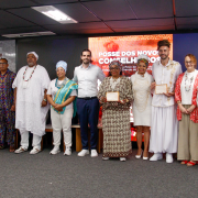 Fotografia mostra um grupo de cerca de doze pessoas adultas em pé, alinhadas lado a lado sobre um palco em um auditório. Algumas seguram certificados retangulares com moldura clara, à frente do corpo. As vestimentas são variadas, incluindo roupas sociais, tecidos estampados e trajes tradicionais com túnicas, colares, lenços e turbantes. Ao fundo há um grande painel digital vermelho com textos em branco, parcialmente legíveis. O piso é cinza, o teto possui luminárias retangulares e há cadeiras distribuídas atrás do grupo.