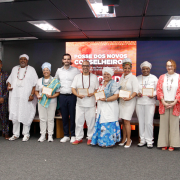 Fotografia mostra um grupo de cerca de treze pessoas adultas em pé, alinhadas lado a lado sobre um palco em um auditório. Todas estão voltadas para a câmera; várias seguram certificados retangulares com moldura clara, posicionados à frente do corpo. As vestimentas são diversas, com roupas sociais, tecidos coloridos, estampas e trajes tradicionais, incluindo colares, turbantes e panos longos. Ao fundo há um grande painel digital vermelho com textos em branco. O piso é cinza, o teto tem luminárias retangulares e há cadeiras dispostas atrás do grupo.