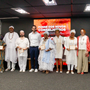 Fotografia mostra um grupo de cerca de treze pessoas adultas em pé, alinhadas lado a lado sobre um palco em um auditório. Todas estão voltadas para a câmera; várias seguram certificados retangulares com moldura clara, posicionados à frente do corpo. As vestimentas são variadas, com roupas sociais, peças claras e trajes tradicionais, incluindo túnicas, tecidos estampados, colares e turbantes. Ao fundo há um grande painel digital vermelho com textos em branco. O piso é cinza, o teto tem luminárias retangulares e há cadeiras dispostas atrás do grupo.