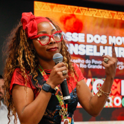 Fotografia mostra uma mulher negra adulta falando ao microfone em um auditório, faz parte da executiva do conselho. Usa roupa vermelha, óculos da mesma cor e faixa vermelha na cabeça, com cabelos longos e claros. Uma das mãos segura o microfone e a outra está levantada, com o dedo indicador apontando para cima. Ao fundo há um painel digital vermelho com textos e grafismos, além de outras pessoas parcialmente visíveis.