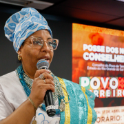 Fotografia mostra uma mulher negra adulta falando ao microfone em um auditório, parte da executiva do conselho. Usa turbante azul com estampa clara, óculos e colares sobre uma roupa clara. Segura o microfone com uma das mãos, próximo ao rosto. Ao fundo há um painel digital vermelho com textos em branco e grafismos. A iluminação é uniforme e o enquadramento destaca a pessoa em primeiro plano.