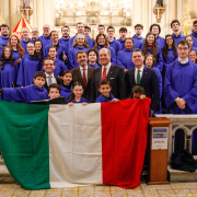 Grupo numeroso posa dentro de uma igreja decorada, com altar ao fundo e detalhes dourados. A maioria usa t&uacute;nicas azuis e segura pastas. Na frente, pessoas exibem uma grande bandeira da It&aacute;lia, nas cores verde, branco e vermelho. Alguns trajes sociais aparecem entre o grupo. O ambiente &eacute; iluminado e possui grades de pedra na parte frontal.