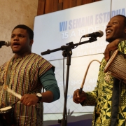Dois homens negros em pé se apresentam diante de microfones. Um bate em tambores pequenos presos ao corpo, usando baquetas; o outro segura um instrumento de percussão com uma mão e uma baqueta na outra. Ambos vestem roupas estampadas. Ao fundo, telão exibe “VII Semana Estadual do Migrante, 15 a 19 de junho de 2026”. Ambiente interno com parede de pedra e madeira.
